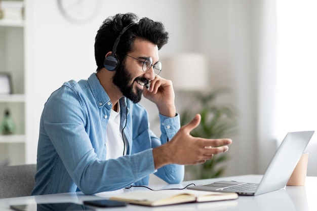 Retrato de homem indiano sorridente no fone de ouvido fazendo videochamada com laptop