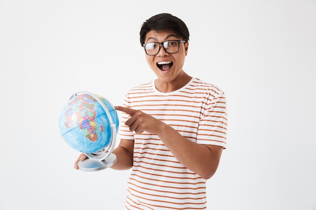 Retrato de homem inteligente estudante asiático vestindo camiseta listrada e óculos, segurando e apontando o dedo para o globo durante a aula, isolado