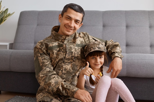 Retrato de homem jovem soldado adulto satisfeito sorridente, vestindo uniforme de camuflagem, voltando para casa depois do exército, passando um tempo com a filha, olhando para a câmera com uma expressão feliz, garoto aparecendo o polegar.