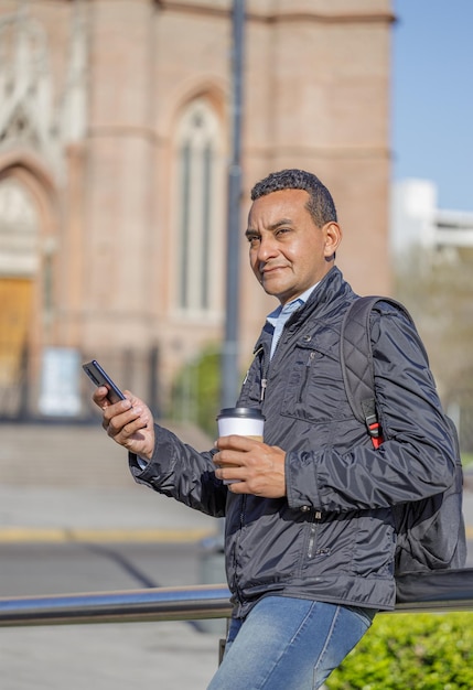 Retrato de homem latino usando seu celular e segurando uma xícara de café descartável