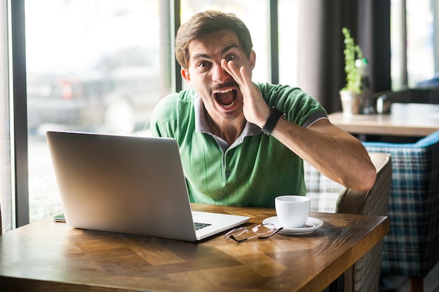 Retrato de homem louco vestindo camiseta verde, sentado com a mão perto da boca e gritando para compartilhar algo enquanto trabalhava on-line no laptop, tiro interno perto da janela grande, fundo do café.