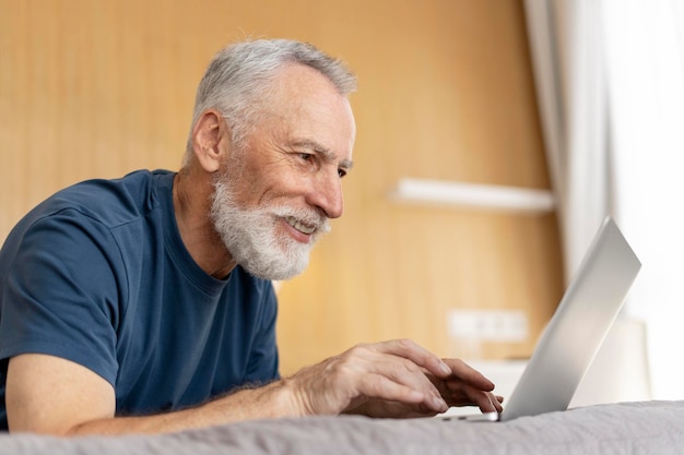 Retrato de homem maduro sorridente confiante com barba usando laptop deitado na cama no hotel Atraente pensionista idoso bem sucedido jogando jogo na sala de estar Conceito de trabalhar em casa