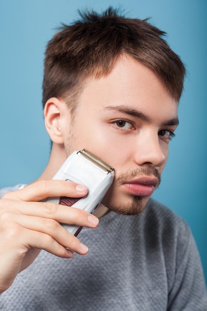 Retrato de homem moreno bonito com cerdas e bigode em suéter casual, removendo pêlos faciais usando depilador para pele lisa, conceito de cuidados de beleza masculino. tiro de estúdio isolado em fundo azul