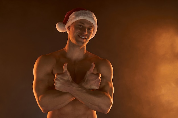 Retrato de homem musculoso com chapéu de Papai Noel de Natal, mãos postas e sorriso no fundo esfumaçado Macho stripper de torso nu, sem camisa