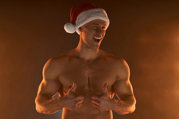 Retrato de homem musculoso com chapéu de Papai Noel de Natal, mostrando o polegar para cima e um sorriso no fundo esfumaçado Macho Shirtless Nu torso stripper Cara de Papai Noel gostoso e sexy Homem sedutor para as férias