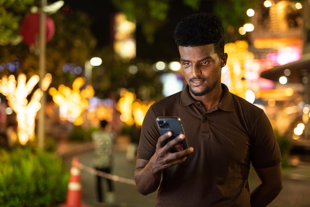 Retrato de homem negro bonito na cidade à noite usando telefone