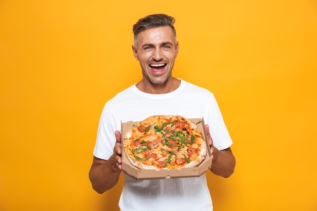 Retrato de homem otimista de 30 anos em uma camiseta branca segurando e comendo pizza em pé isolado no amarelo