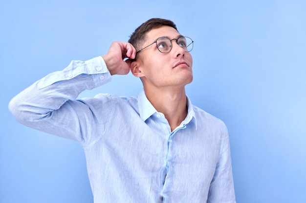 Retrato de homem pensativo de óculos e camisa toca sua cabeça olha para o canto superior e pensa que toma uma decisão com cara de dúvida isolada em estúdio azul com espaço de cópia