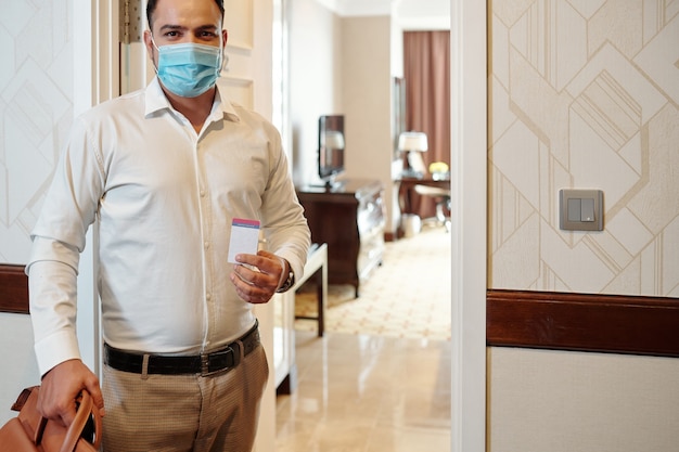 Retrato de homem positivo com máscara médica em pé no quarto de hotel com mala e chave digital