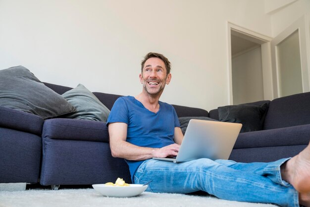 Retrato de homem rindo com laptop sentado no chão da sala de estar