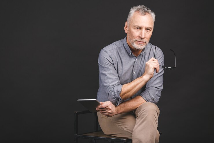 Foto retrato de homem sênior aposentado, segurando na mão um tablet digital enquanto está sentado, segurando os óculos.