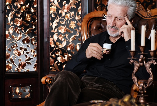 Retrato de homem sênior bebendo em casa enquanto está sentado na cadeira vintage