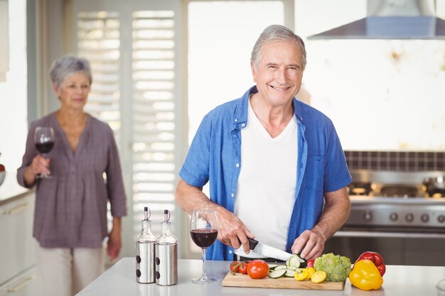 Retrato de homem sênior cortar legumes com a esposa em segundo plano