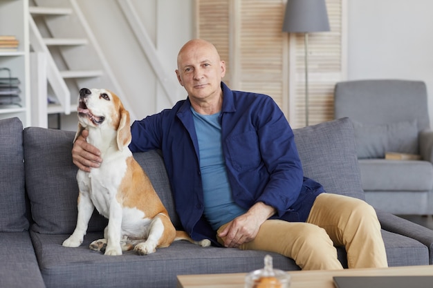 Retrato de homem sênior moderno com cachorro sentado no sofá, posando no interior da casa