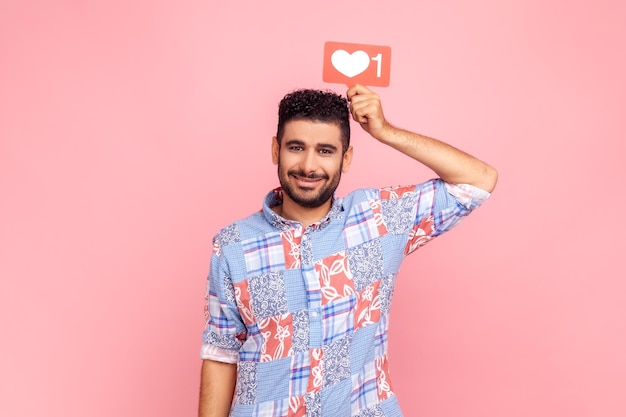 Retrato de homem sorridente com barba e cabelo escuro segurando como ícone acima da cabeça, olhando para a câmera com expressão facial satisfeita, mídia social. Tiro de estúdio interior isolado no fundo rosa.