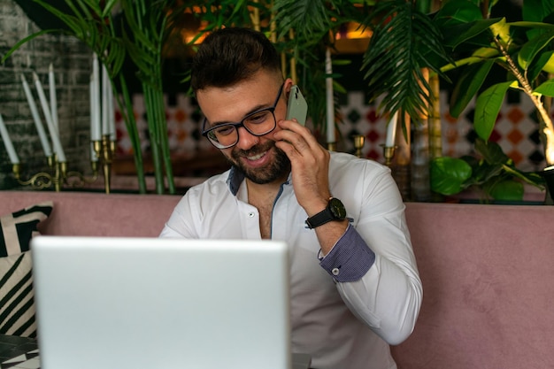 Retrato de homem sorridente com barba sentado com laptop, falando ao telefone