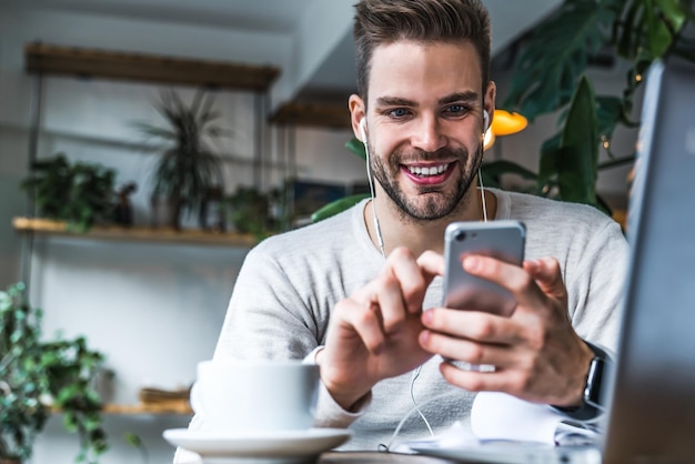 Retrato de homem sorridente com fones de ouvido sentado no café e usando telefone celular