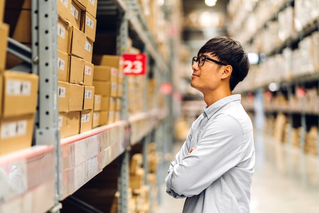 Retrato de homem sorridente trabalhador gerente asiático em pé e detalhes do pedido, verificando mercadorias e suprimentos nas prateleiras com fundo de mercadorias em warehouse.