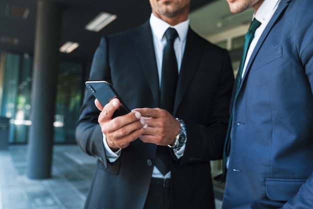 Retrato de homens de negócios caucasianos parceiros vestidos com um terno formal em pé do lado de fora do centro de trabalho e usando o celular juntos durante a reunião de trabalho