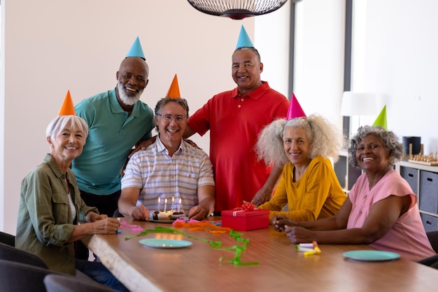 Retrato de idosos multirraciais alegres usando chapéus de festa enquanto comemorava o aniversário do amigo. Casa de repouso, bolo, festa, diversão, inalterado, união, apoio, moradia assistida, aposentadoria.