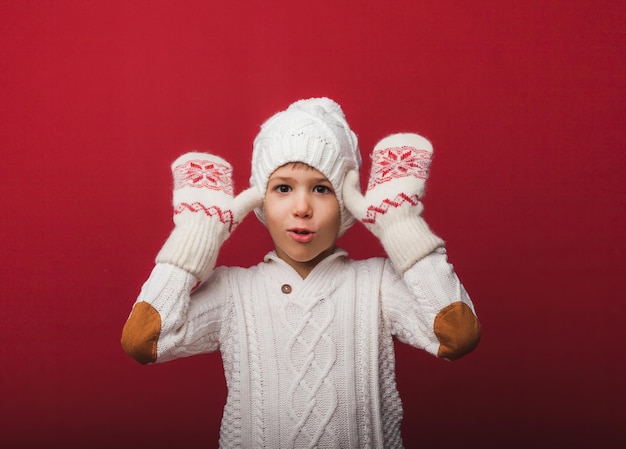 Retrato de inverno de um menino feliz e alegre em um chapéu de malha e suéter se divertindo em um fundo vermelho o menino olha para as palmas das mãos