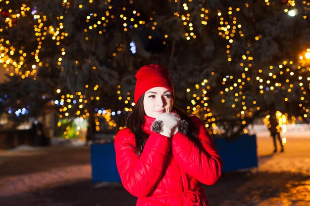 Retrato de inverno quente de uma jovem adorável com longos cabelos escuros com casaco vermelho e chapéu, segurando as mãos em luvas de lã quentes perto do rosto. Árvore de Natal da cidade grande com luzes no fundo