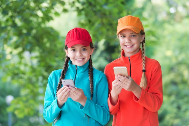 Retrato de irmãs felizes com smartphones crianças se divertem Duas garotas hipster sorridentes em roupas esportivas crianças despreocupadas posando ao ar livre Modelos positivos Roupa casual de descolados