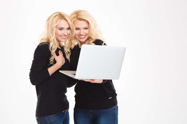 Retrato de irmãs gêmeas sorridentes usando um computador laptop isolado em um fundo branco