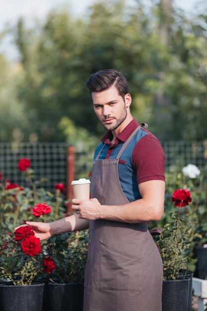 Retrato de jardineiro bonito em avental segurando xícara de café descartável no jardim