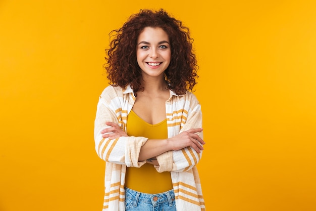 Retrato de jovem 20 anos com cabelo encaracolado sorrindo e em pé com os braços cruzados, isolado no amarelo