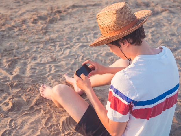 Retrato de jovem adolescente brincando com seu telefone na praia do mar com chapéu de verão