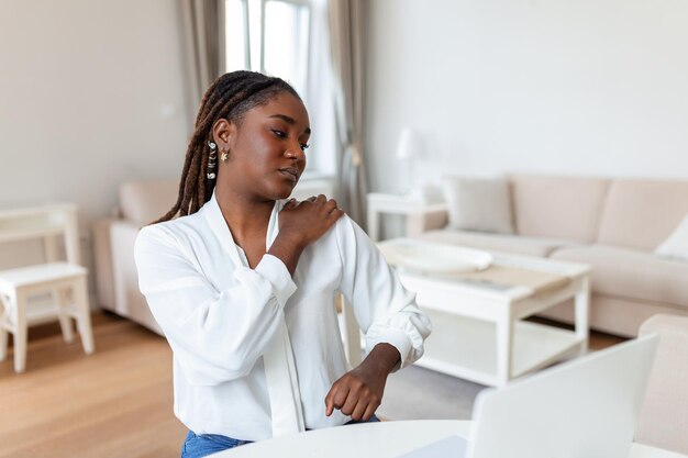 Retrato de jovem africana estressada sentada na mesa de escritório em casa na frente do laptop tocando ombro dolorido com expressão de dor sofrendo de dor no ombro depois de trabalhar no pc