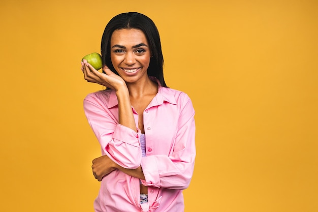Foto retrato de jovem afro-americana demonstrando sorriso saudável segurando maçã verde recomendando serviço de clareamento dental isolado sobre fundo amarelo