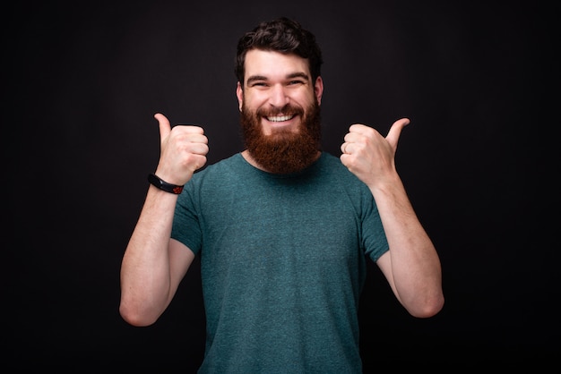 Retrato de jovem alegre com barba aparecendo polegares em pé sobre fundo escuro