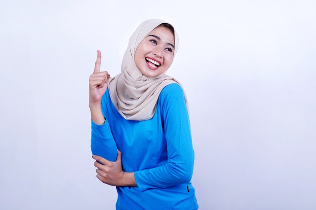 Retrato de jovem alegre com camiseta azul usando hijab, sentindo-se feliz e apontando para o lado esquerdo