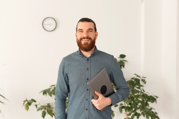 Retrato de jovem alegre em pé casual no escritório e segurando o laptop