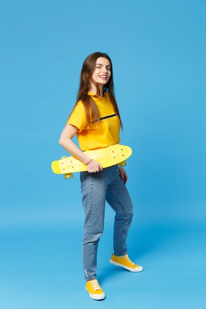 Retrato de jovem alegre em roupas casuais vívidas em pé, olhando a câmera, segurando o skate amarelo isolado no fundo da parede azul no estúdio. Conceito de estilo de vida de pessoas. Mock up espaço de cópia.