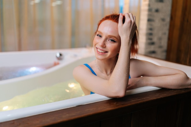 Retrato de jovem alegre em traje de banho relaxado sentado na banheira com hidromassagem no salão spa de luxo olhando para a câmera Fêmea feliz deitada na banheira