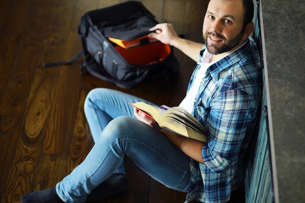 Retrato de jovem alegre lendo livro enquanto está sentado no chão de sua sala de estar. aluno segurando e lendo o livro.