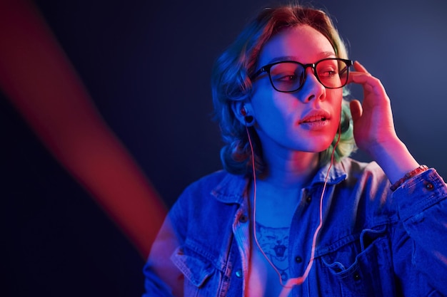 Retrato de jovem alternativa em óculos com cabelo verde em luz neon vermelha e azul no estúdio que ouve música em fones de ouvido