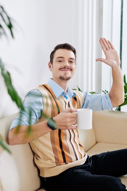 Retrato de jovem animado e feliz com uma xícara de café, acenando com a mão e olhando para a câmera