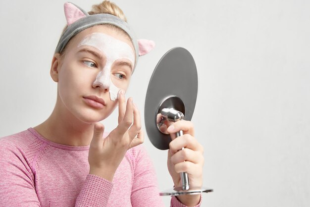 Retrato de jovem aplicando máscara facial cosmética de argila branca olhando no espelho isolado na parede branca, cuidados faciais em casa, rotina feminina de manhã e noite