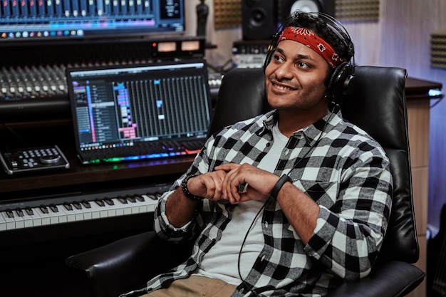Retrato de jovem artista masculino feliz em fones de ouvido sorrindo à parte enquanto está sentado na gravação