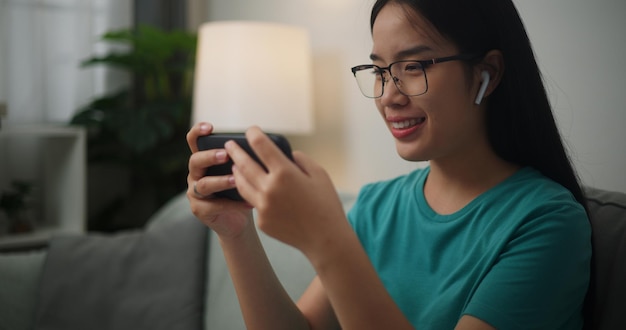 Retrato de jovem asiática usando óculos e fones de ouvido gosta de jogar jogos online em seu smartphone no sofá na sala de estarConceito de estilo de vida do jogador