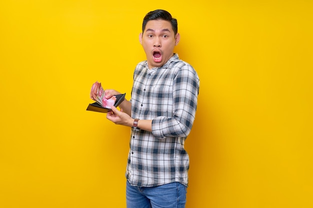 Retrato de jovem asiático chocado vestindo camisa xadrez segurando uma carteira cheia de dinheiro em notas de rupia na mão isoladas no conceito de estilo de vida de pessoas de fundo amarelo
