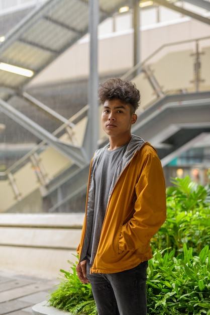 Retrato de jovem asiático com cabelo encaracolado na cidade ao ar livre com chuva