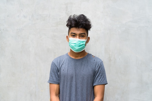 Retrato de jovem asiático com cabelo encaracolado usando máscara para proteção contra surto de coronavírus na parede de concreto