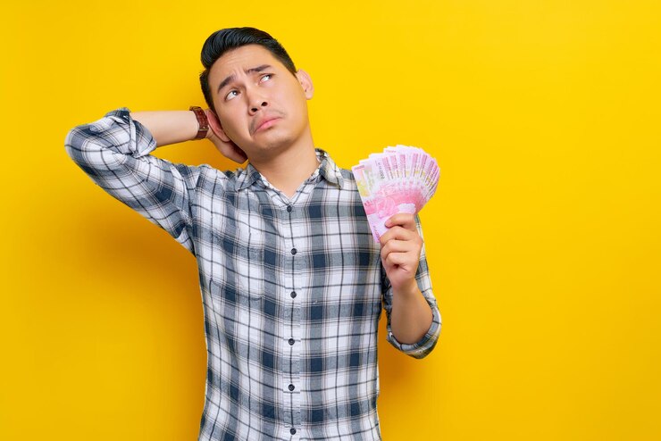 Foto retrato de jovem asiático confuso vestindo camisa xadrez segurando um leque de dinheiro em notas de rupia e olhando para o espaço vazio isolado no conceito de estilo de vida de pessoas de fundo amarelo