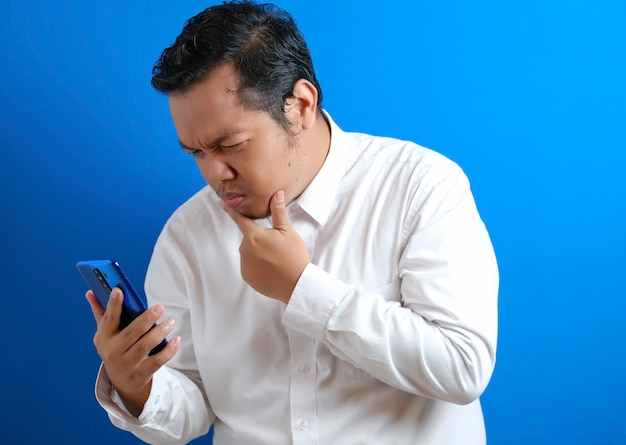 Retrato de jovem asiático em camisa branca casual tentando ler atentamente as notícias em seu telefone inteligente, expressão facial curiosa