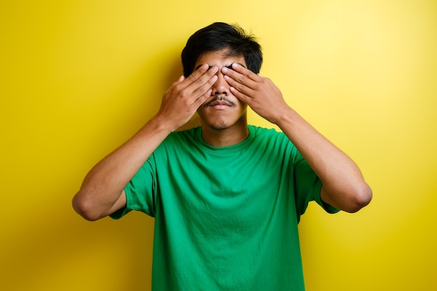 Retrato de jovem asiático em uma camiseta verde, cobrindo os olhos com as duas mãos contra um fundo amarelo